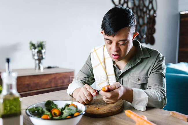 Підліток з синдромом Дауна сидить за столом і ріже овочі під час приготування салату на обід вдома — стокове фото