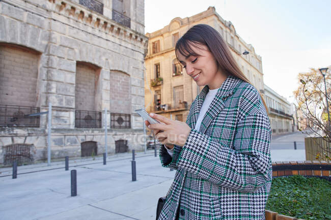 Cheerful young female in stylish houndstooth coat using mobile phone while standing on urban square — Stock Photo