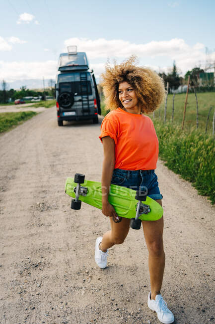 Heureuse femme afro-américaine portant longboard et regardant loin avec sourire tout en marchant sur la route de campagne près de van en été — Photo de stock