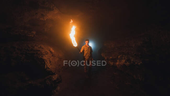 Joven espeleólogo masculino con antorcha encendida de pie en la oscura cueva rocosa estrecha mientras explora el entorno subterráneo mirando a la cámara - foto de stock
