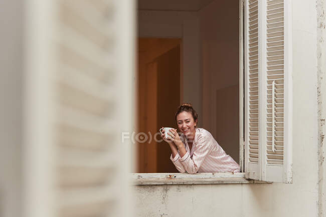 Tranquil hembra en pijama inclinada en la ventana con taza de café de la mañana y mirando a la cámara - foto de stock
