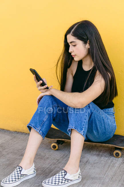 Junge Skaterin surft mit Handy im Internet und sitzt tagsüber auf Skateboard auf Gehweg — Stockfoto