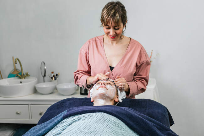 Estetista femminile che applica il detergente facciale sul viso del cliente femminile durante il trattamento della pelle nel salone di bellezza — Foto stock