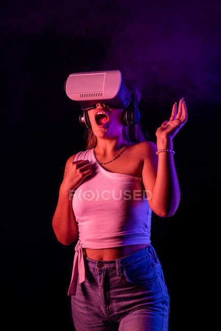 Donna irriconoscibile in VR auricolare che interagisce con la realtà virtuale mentre in piedi in studio scuro con vapore e illuminazione al neon blu e rosa — Foto stock