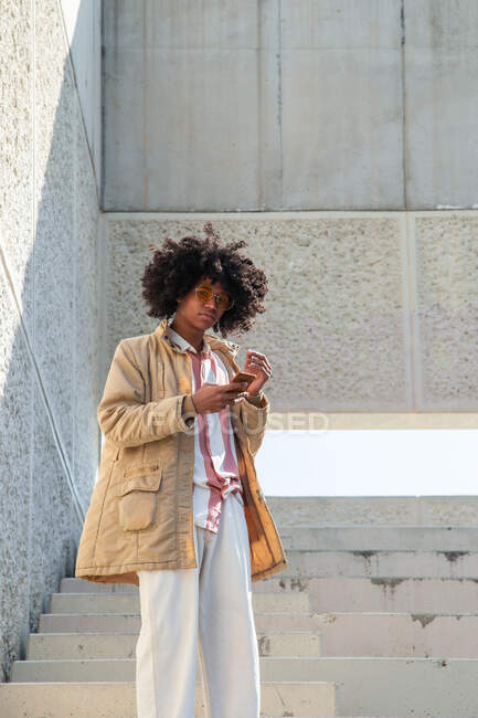 Bajo ángulo de hombre étnico en abrigo vintage con mensajes de texto de peinado afro en el teléfono celular en la escalera urbana - foto de stock