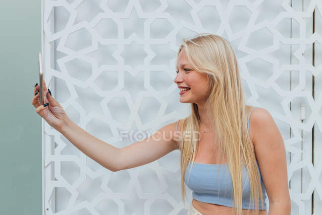 Vue latérale d'une femme joyeuse avec des cheveux blonds se tirant dessus sur un téléphone portable dans la rue de la ville — Photo de stock