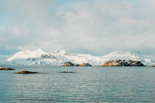 Кам'яні острівці, розташовані в хвилястому морі біля снігового гірського хребта проти хмарного неба взимку на Лофотенських островах, Норвегія. — стокове фото
