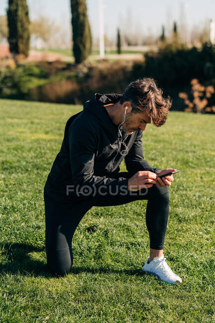 Atleta barbudo em roupas esportivas e fones de ouvido mensagens de texto no celular enquanto se agacha no prado na cidade — Fotografia de Stock