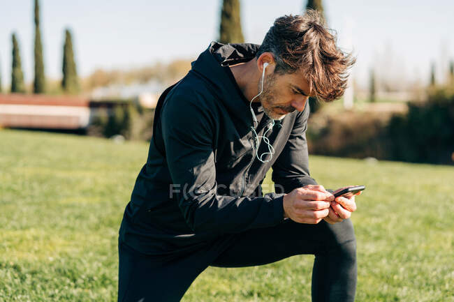 Atleta masculino barbudo en ropa deportiva y auriculares mensajería de texto en el teléfono celular mientras se agacha en el prado de la ciudad - foto de stock