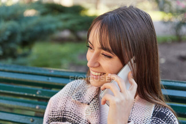 Desde arriba, una mujer milenaria sonriente moderna con un elegante atuendo de primavera sentada en el banco y respondiendo a una llamada telefónica mientras descansa en la calle urbana mirando hacia otro lado. - foto de stock