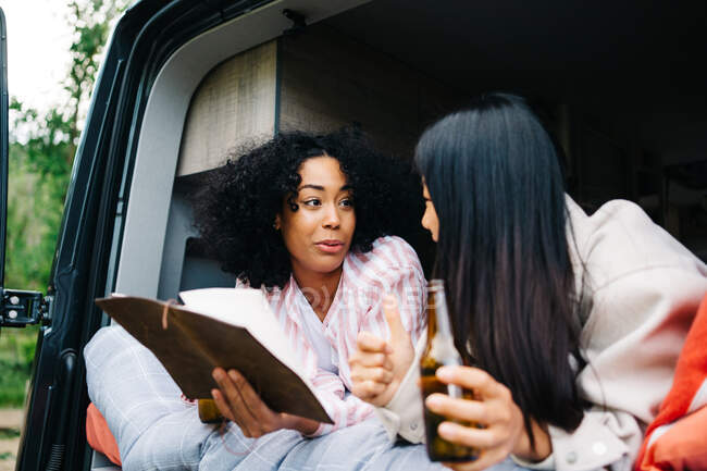 Jovens mulheres multirraciais alegres bebendo cerveja e discutindo a rota de viagem com o mapa enquanto relaxam juntas em van campista durante a viagem de verão — Fotografia de Stock