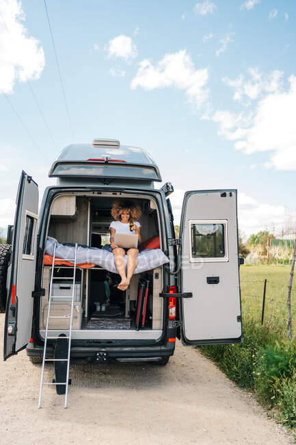 Young African American female traveler with curly hair drinking beer and watching movie on laptop while resting inside camper van during summer holidays — Stock Photo