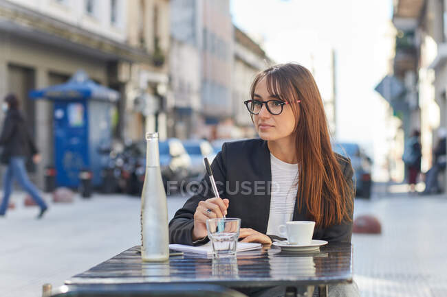 Позитивная молодая деловая леди в элегантном костюме и очках делает заметки в блокноте, сидя за столом в уличном кафе в городе, отворачиваясь — стоковое фото