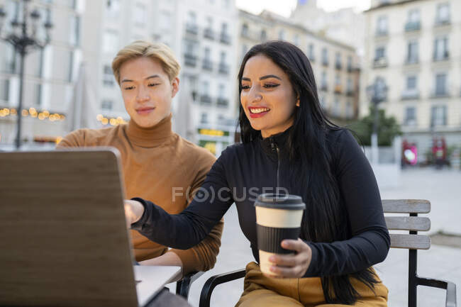 Alegre freelancer multirracial sentado en la mesa con el ordenador portátil y trabajando en el proyecto remoto juntos en la cafetería al aire libre - foto de stock