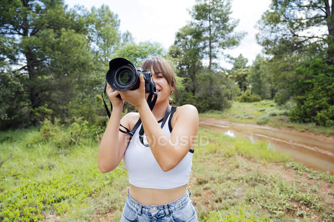 Задоволена подорожуюча жінка-фотограф фотографує професійну камеру під час відпустки в лісі — стокове фото