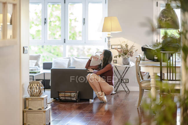 Side view of slender female ballet dancer in bodysuit and pointe shoes balancing on tiptoes near couch in living room at home — Stock Photo