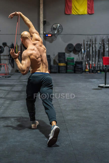 Back view of unrecognizable shirtless man stretching elastic band with arms and lunging during functional workout in gym — Stock Photo