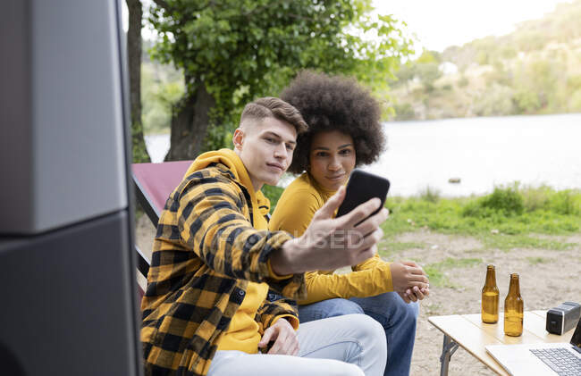 Young man taking selfie with black girlfriend while sitting outside van on summer day in countryside — Stock Photo