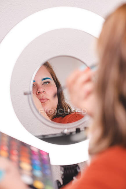 Back view of plump female using brush to apply makeup near ring light in studio — Stock Photo