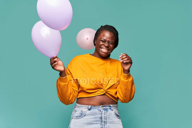 Emocionada mujer afroamericana en jeans y top amarillo de pie cerca de globos cayendo sobre fondo azul en el estudio - foto de stock