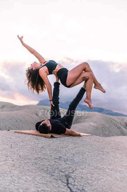 Vista lateral do casal gracioso fazendo acroyga no fundo do céu por do sol nas montanhas — Fotografia de Stock