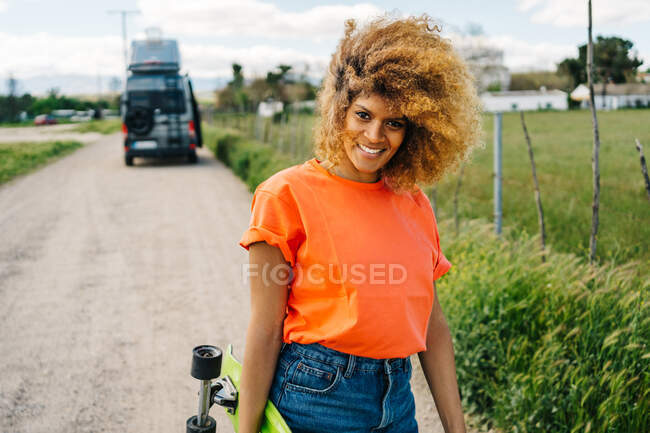 Mulher afro-americana feliz carregando longboard e olhando para a câmera com sorriso enquanto caminhava na estrada rural perto de van no verão — Fotografia de Stock