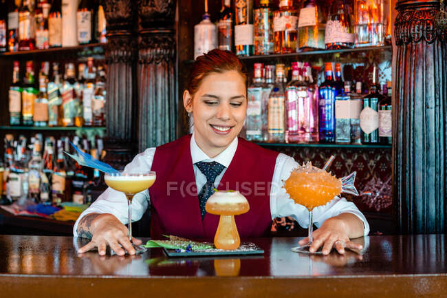 Cantinera sonriente parada en el mostrador del bar con diferentes tipos de bebidas alcohólicas servidas en vasos de cóctel creativos en forma de champiñón y pescado - foto de stock