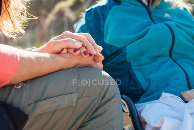 Cultiver femme soignante méconnaissable tenant tendrement la main de la femme âgée en fauteuil roulant sur une journée ensoleillée — Photo de stock