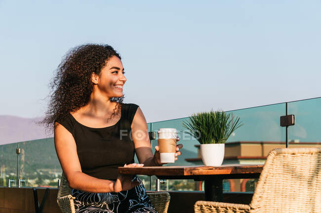Heureux jeune fille bouclée hispanique boire du café à emporter et profiter de la journée d'été tout en étant assis à table sur la terrasse du café — Photo de stock