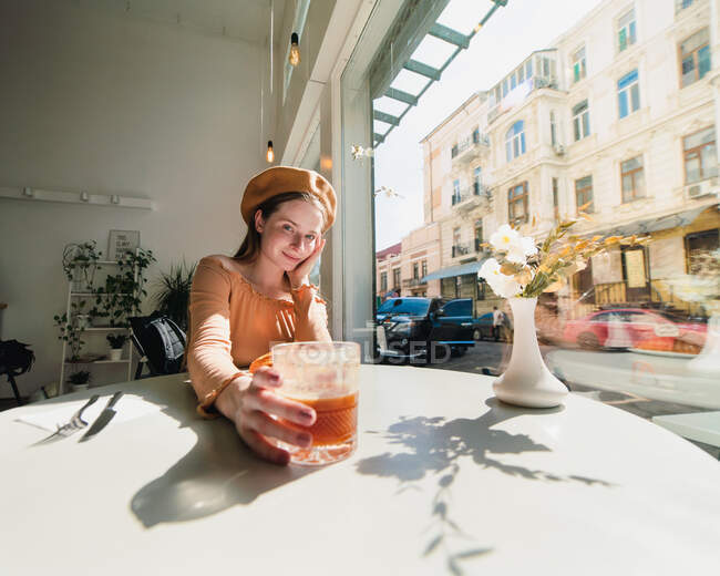 Hembra francesa en boina sentada a la mesa en la cafetería con vaso aromático de café y croissant recién horneado - foto de stock