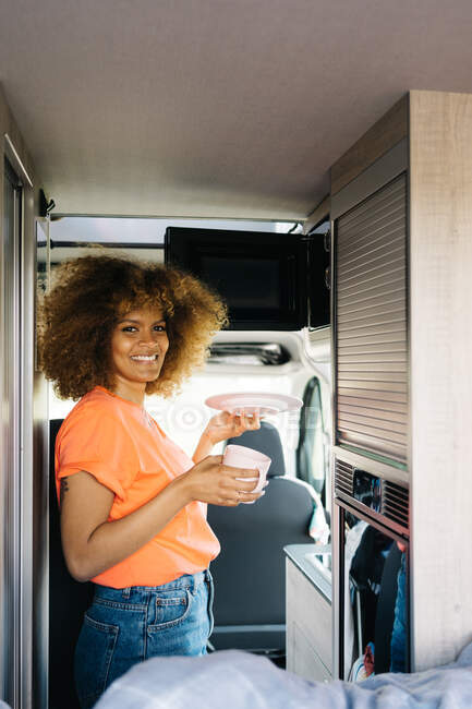 Vue latérale du voyageur féminin avec une plaque de mise en cheveux bouclés dans un four à micro-ondes tout en chauffant les aliments à l'intérieur de la caravane moderne — Photo de stock