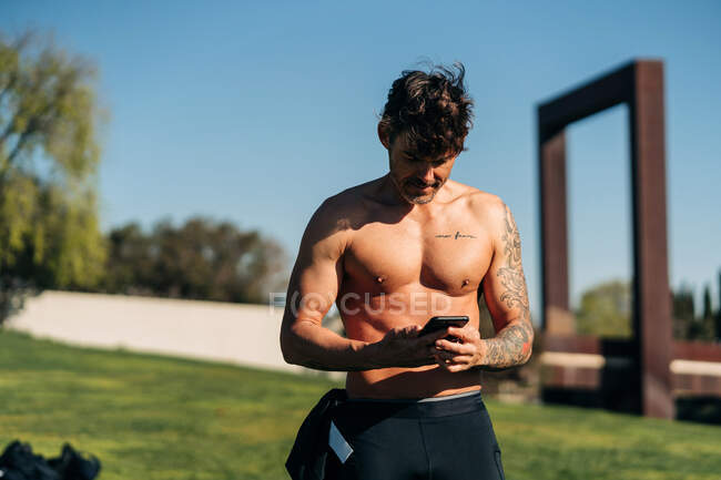 Cheerful male athlete with naked torso browsing cellphone after training on sunny day — Stock Photo