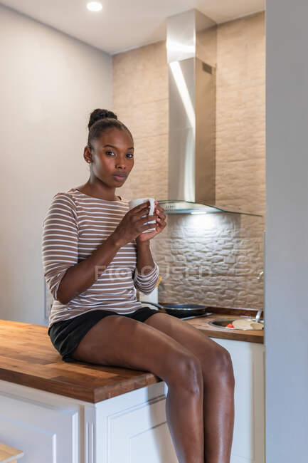 Vista lateral de una joven afroamericana soñadora con taza de bebida caliente sentada en la mesa y mirando a la cámara en el interior - foto de stock