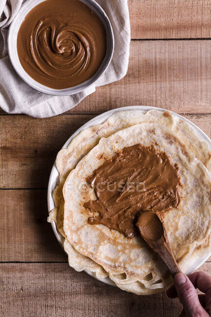 From above anonymous person using spoon to spread sweet nut butter of delicious crepes over lumber table — Stock Photo