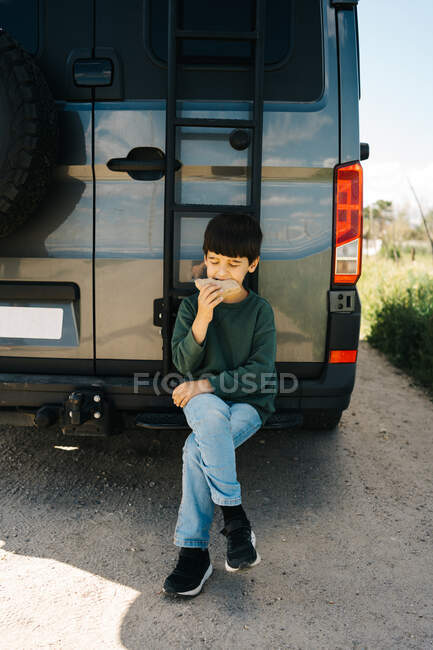 Cute little traveler biting tasty sandwich and looking away while sitting on ladder of modern RV on summer day in countryside — Stock Photo