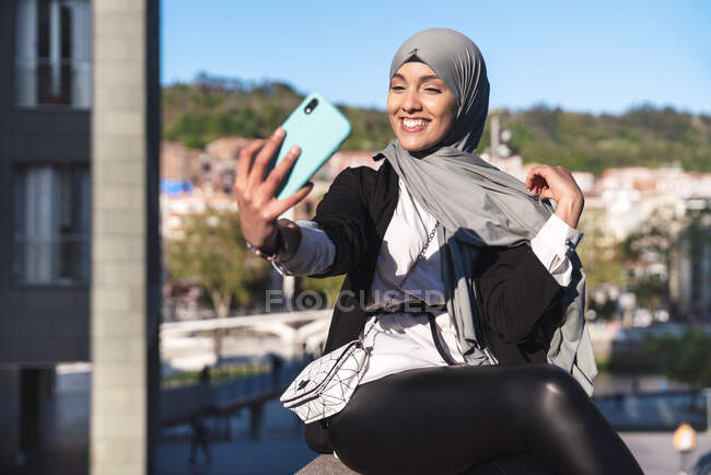 Positive Muslimin im Hidschab macht an sonnigem Tag Selbstporträt auf Smartphone in der Stadt — Stockfoto