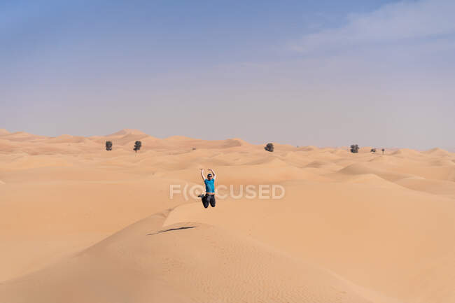 Man in casual clothes jumping on sandy dune against desert during travel in Emirates and stretching arms — Stock Photo