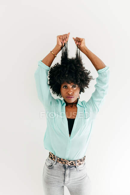 Playful young African American female in trendy outfit having fun touching afro hair on white background — Stock Photo