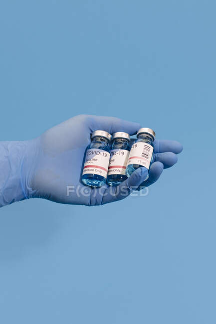Обрізана нерозпізнана рука лікаря, що тримає кожух вакцини проти коронавірусу на синьому фоні — стокове фото