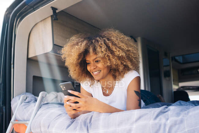 Mulher afro-americana feliz com cabelo encaracolado sorrindo e navegando celular enquanto descansa na cama em caravana durante a viagem de carro — Fotografia de Stock