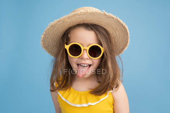Playful little girl in bright yellow swimsuit and sunglasses with straw hat having fun and showing tongue while enjoying summertime against blue background — Stock Photo