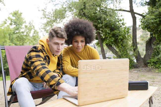 Jovem homem e mulher diverso em roupas casuais sorrindo e navegando netbook enquanto sentado perto de van durante a viagem na natureza — Fotografia de Stock
