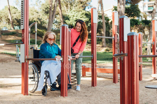 Contenu femme adulte aidant mère aînée en fauteuil roulant faire des exercices sur le terrain de sport pendant la séance de réadaptation — Photo de stock