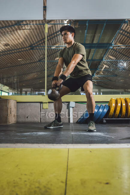 Азиатский мужчина тренировки плечи и руки с тяжелыми гирями в тренажерном зале во время функциональной тренировки и глядя в сторону — стоковое фото