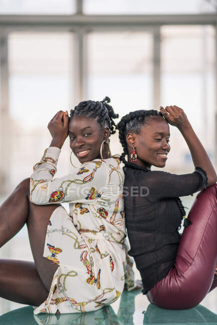 Contenido elegante damas afroamericanas sentadas cerca de nuevo a la espalda y mirando a la cámara con una sonrisa pensativa - foto de stock