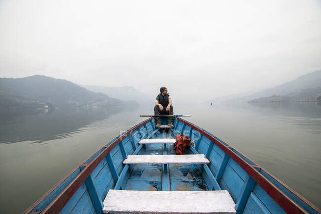 Explorador masculino flutuando em barco de madeira azul no lago calmo na manhã nebulosa durante as férias no Nepal — Fotografia de Stock