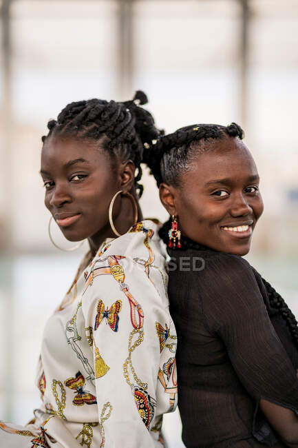 Zufriedene Afroamerikanerinnen, die Rücken an Rücken stehen und bei hellem Tag mit nachdenklichem Lächeln in die Kamera schauen — Stockfoto