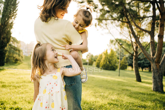 Симпатична дівчина дошкільника посміхається і дивиться геть, коли матуся з маленькою сестрою на руках під час літнього дня разом у зеленому парку — стокове фото