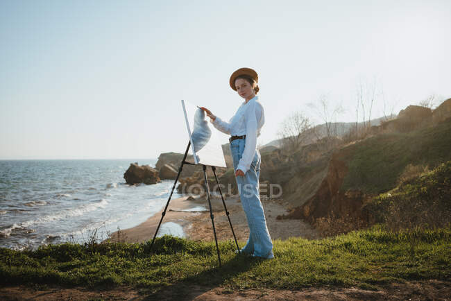 Vue latérale de la jeune femme en vêtements élégants et béret debout sur la côte herbeuse près du sable et de l'océan dans une journée ensoleillée tout en dessinant une image avec pinceau sur toile sur chevalet — Photo de stock