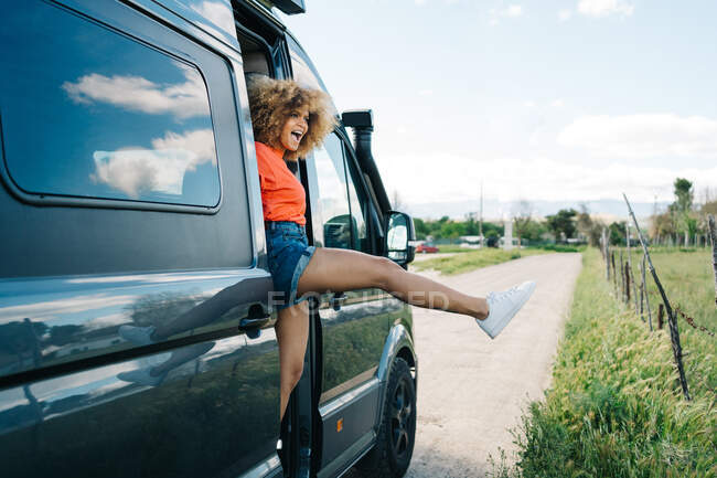Здивована афро-американська жінка з кучерявим волоссям захоплюється сільською місцевістю, коли він стискає ногу з дверей фургона під час дорожньої подорожі влітку. — стокове фото